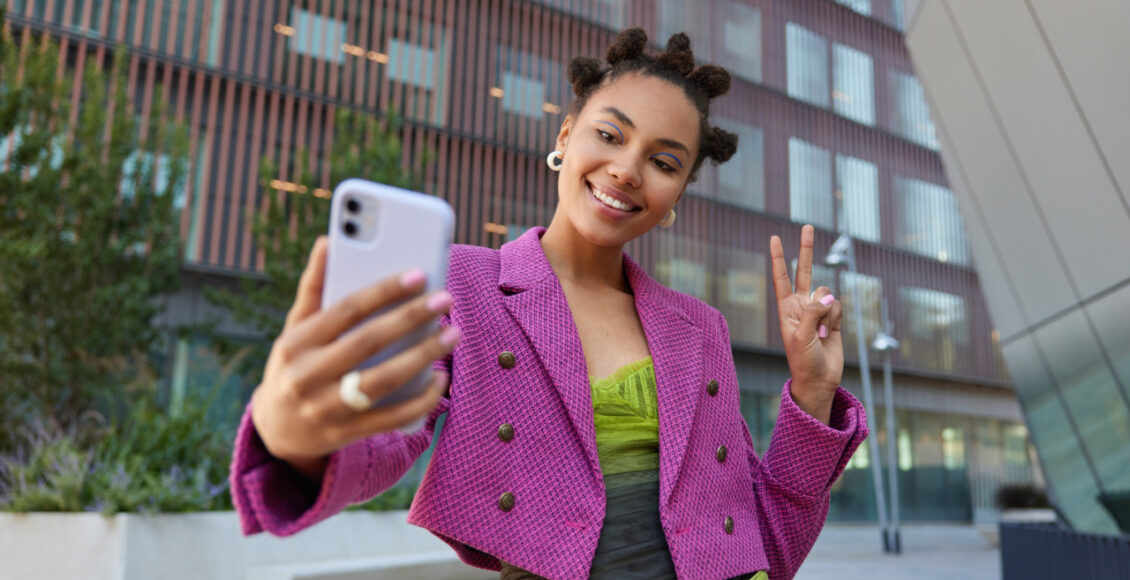 Attractive Girl In Fashionable Outfit Creats Influence Content Shows Peace Sign At Smartphone Front Camera Poses For Selfie Smiles Gladfully Poses At Urban Place Spends Vacation At Big City.