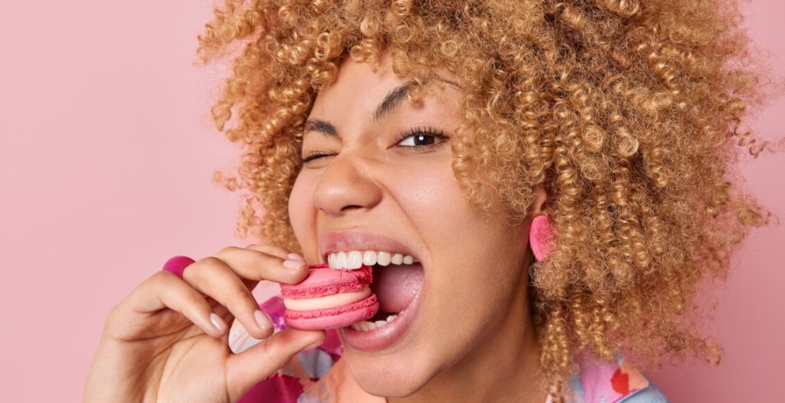 Close Up Shot Of Pretty Curly Haired Woman Eats Delicious French Macaroon Enjoys Dessert Bakery Sweet Yummy Food Keeps Mouth Widely Opened Poses Against Pink Background. Unhealthy Eating Concept