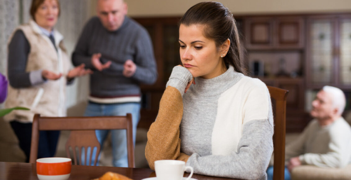 Woman Sitting At Table, Unhappy Family Quarrelling On Backgroud