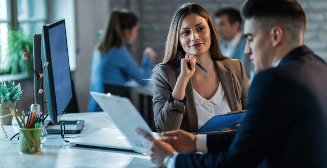 Happy Businesswoman Talking With A Colleague While Going Together Through Paperwork.