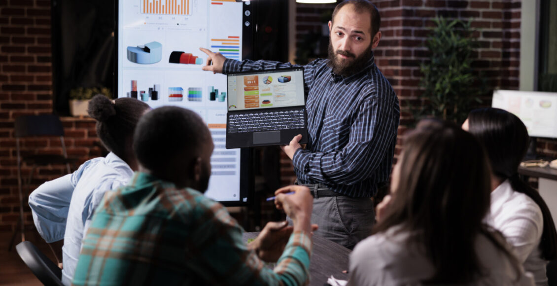 Startup Entrepreneur Holding Laptop With Sales Presentation Charts Pointing At Wall Screen Tv