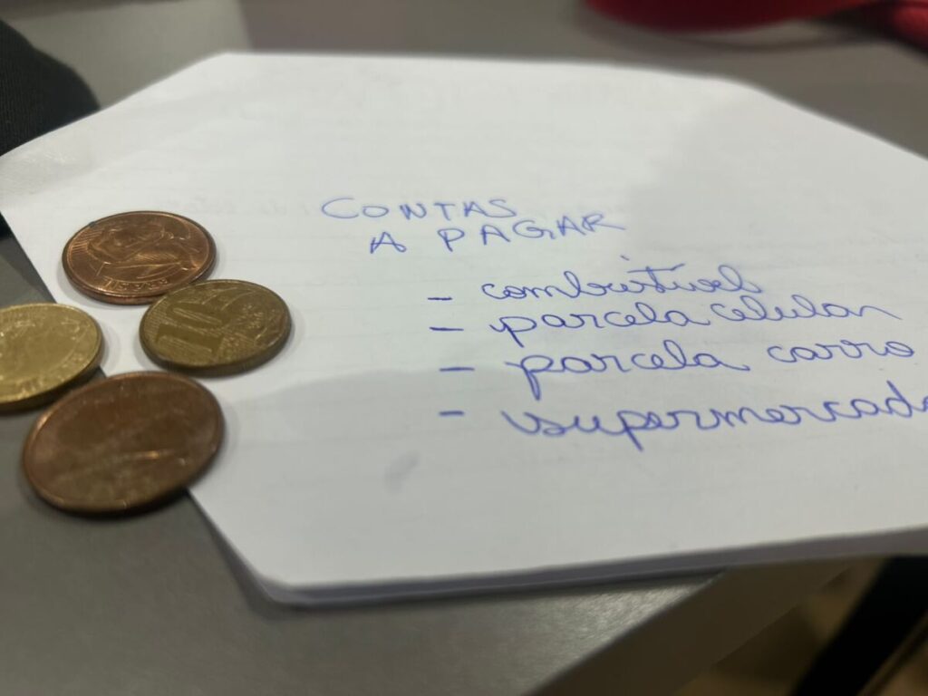 Na imagem, há duas moedas de 5 centavos e duas moedas de 10 centavos. Ao lado, um papel escrito "contas a pagar", com tópicos escritos combustivel, parcela celular, parcela carro e supermercado. Imagem ilustrativa.