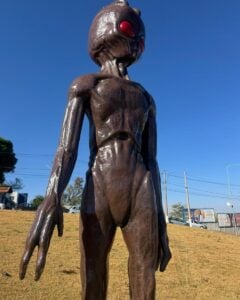 a imagem mostra um et de escultura. atrás, o cenário de uma grama amarelada, o céu azul e carros e estabelecimentos