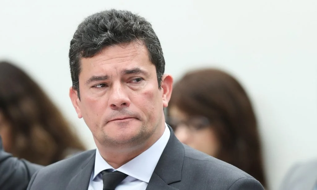 na foto, o ex-ministro apresenta um semblante serio e há um fundo branco e uma mulher desfocada atras. Moro está bem próximo no enquadramento, mostrando seu rosto e parte do ombro